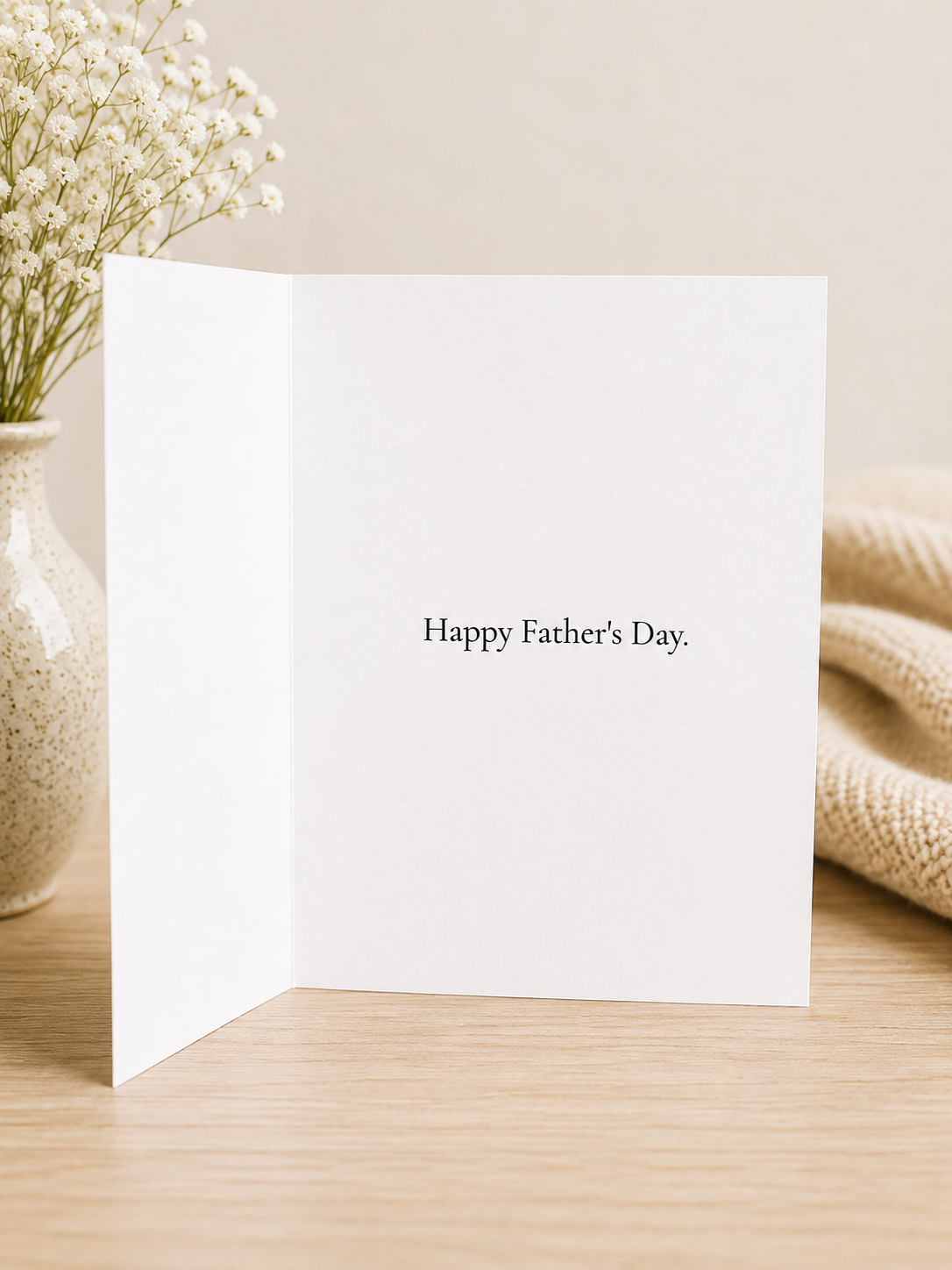 White card with 'Happy Father's Day' text on a wooden surface with a vase of flowers and beige fabric.