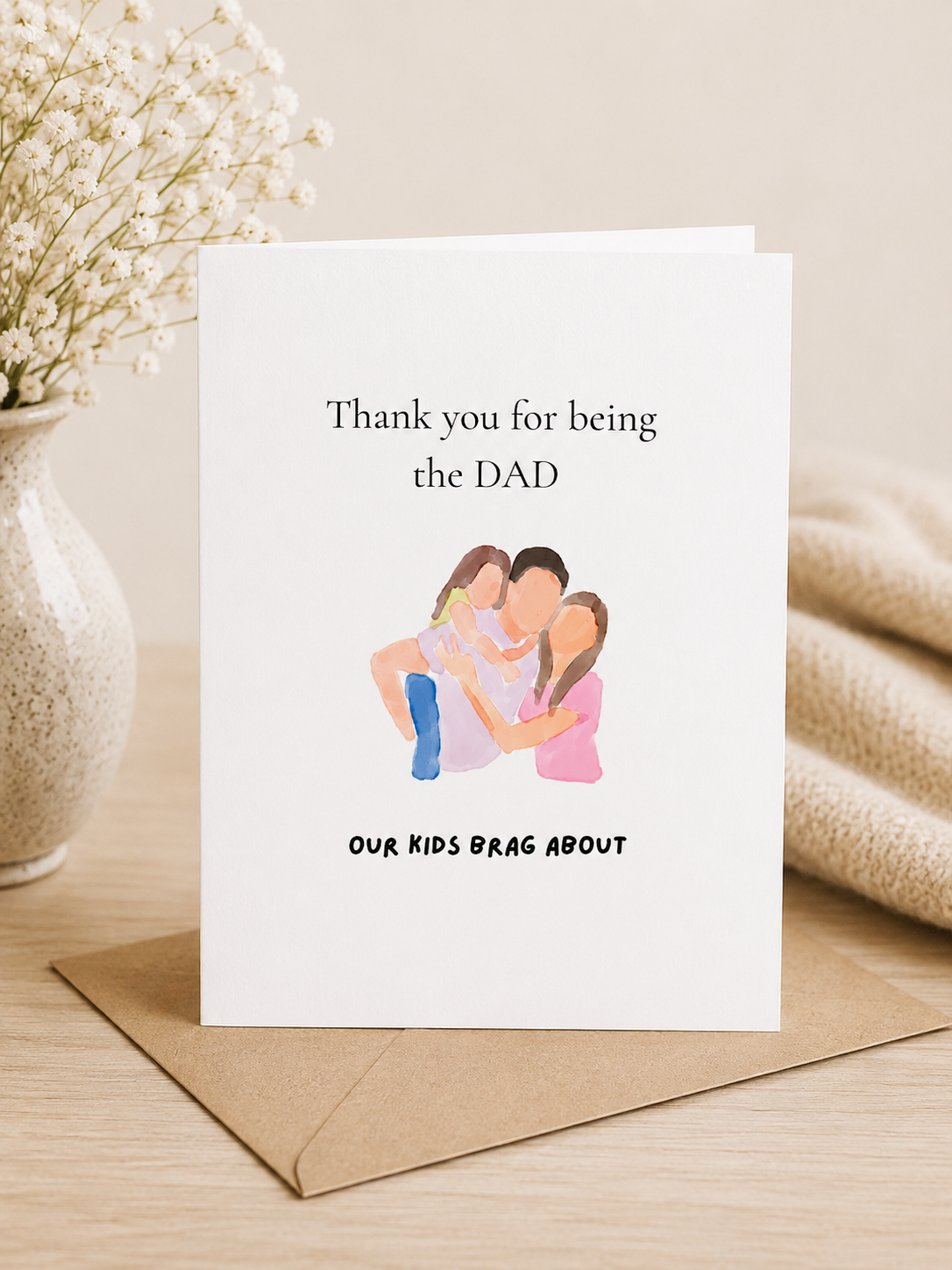 Greeting card with illustration of a family and text on a wooden surface with a vase and blanket in the background