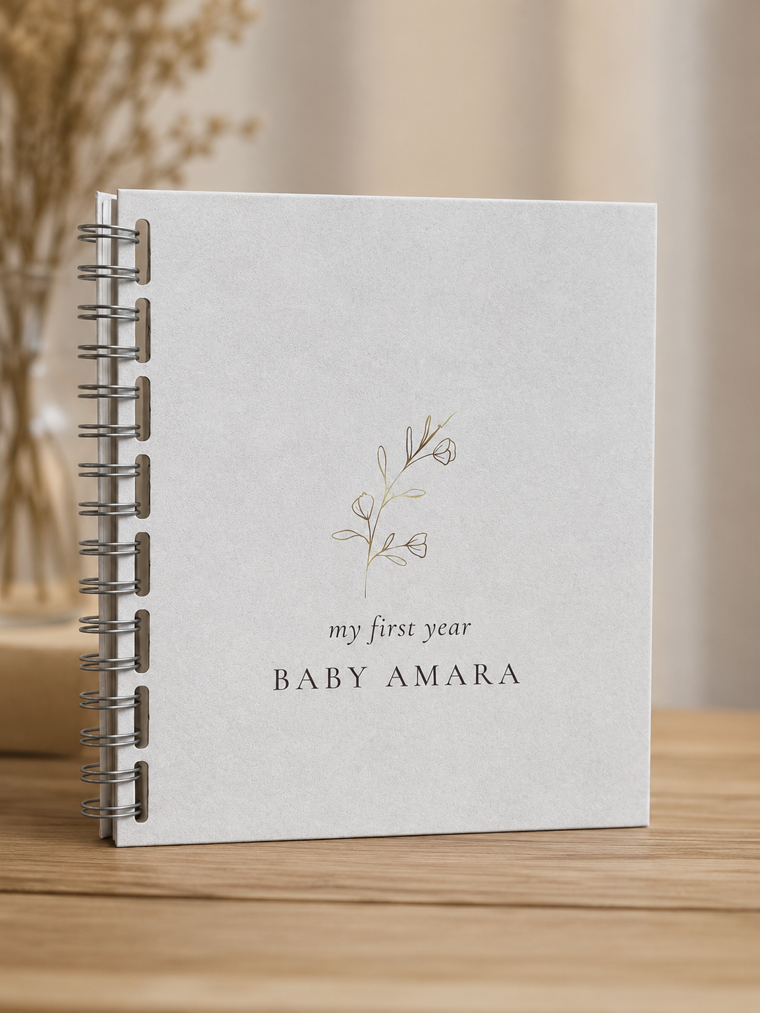 A personalized spiral-bound baby keepsake notebook standing upright on a wooden surface. The notebook cover is a textured off-white with a delicate gold floral illustration and the text 'my first year 