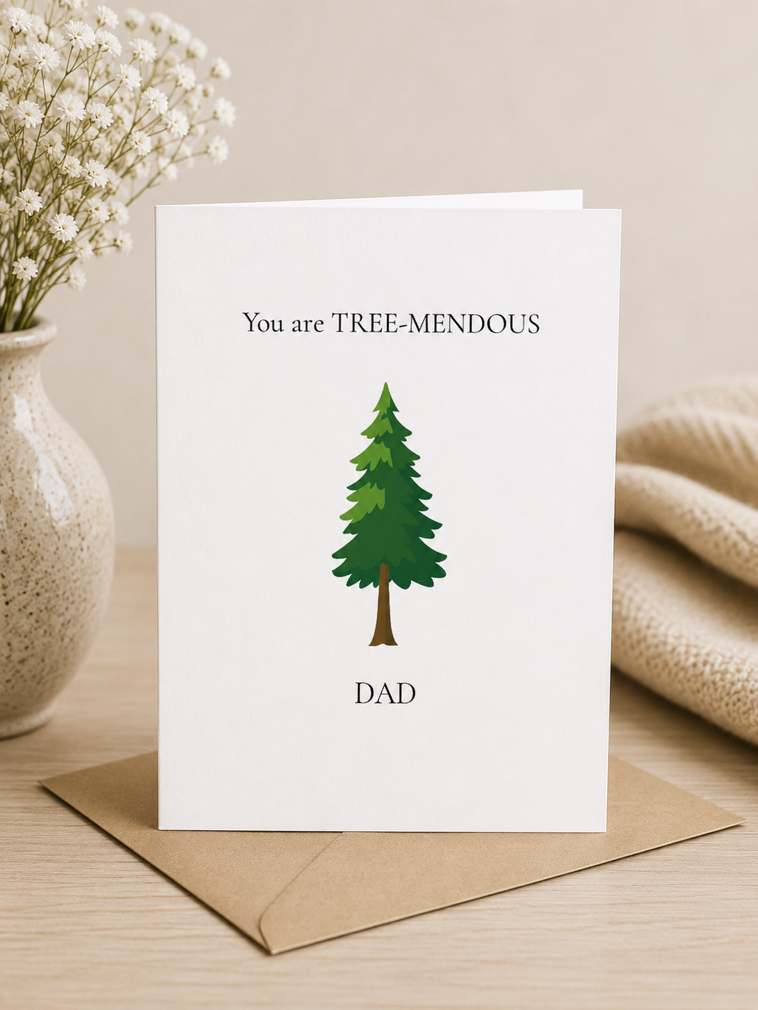 Greeting card with a tree illustration and text on a wooden surface with a vase of flowers and blanket.