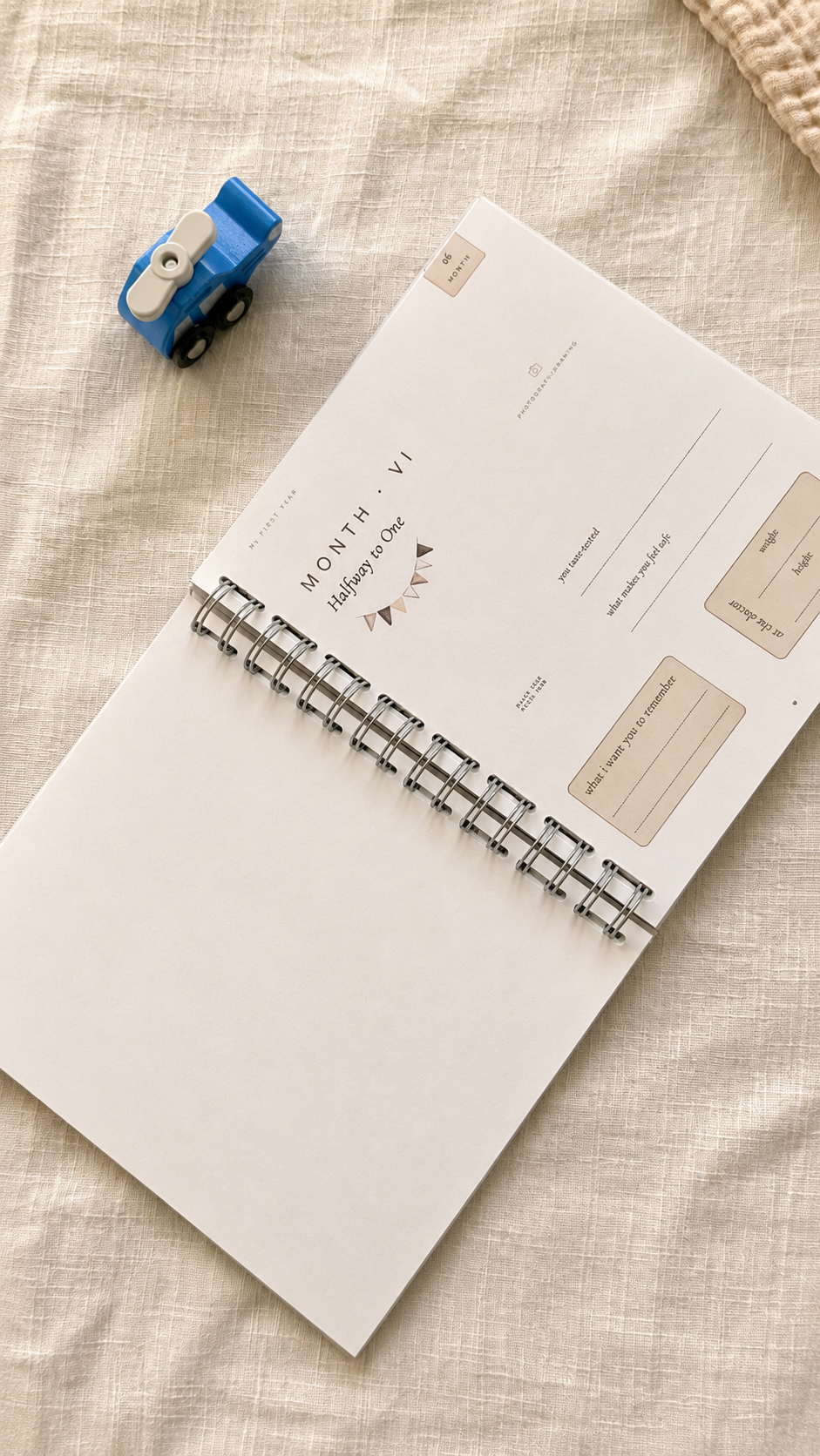 A spiral-bound planner or notebook with a minimalist cream cover lies on a textured beige fabric surface, alongside a small blue toy truck.