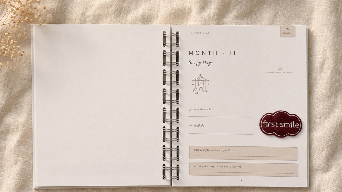An open spiral-bound journal or planner spread on a beige fabric background. The right page shows printed monthly planning sections, including a small illustration of a hanging crib and a dark red oval label reading 'first smile'.